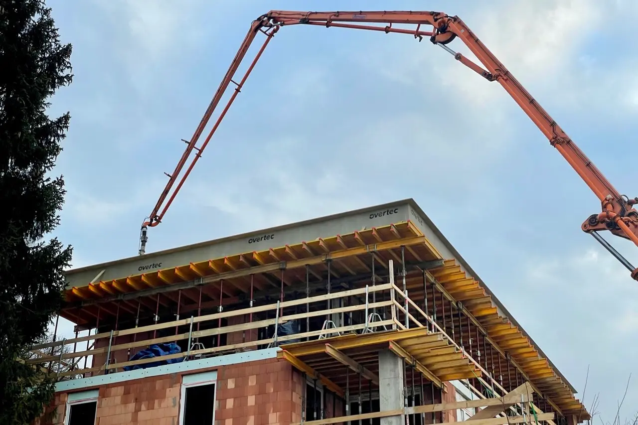 A large orange concrete pump arm reaches over a building construction site featuring yellow scaffolding and Overtec formwork elements.