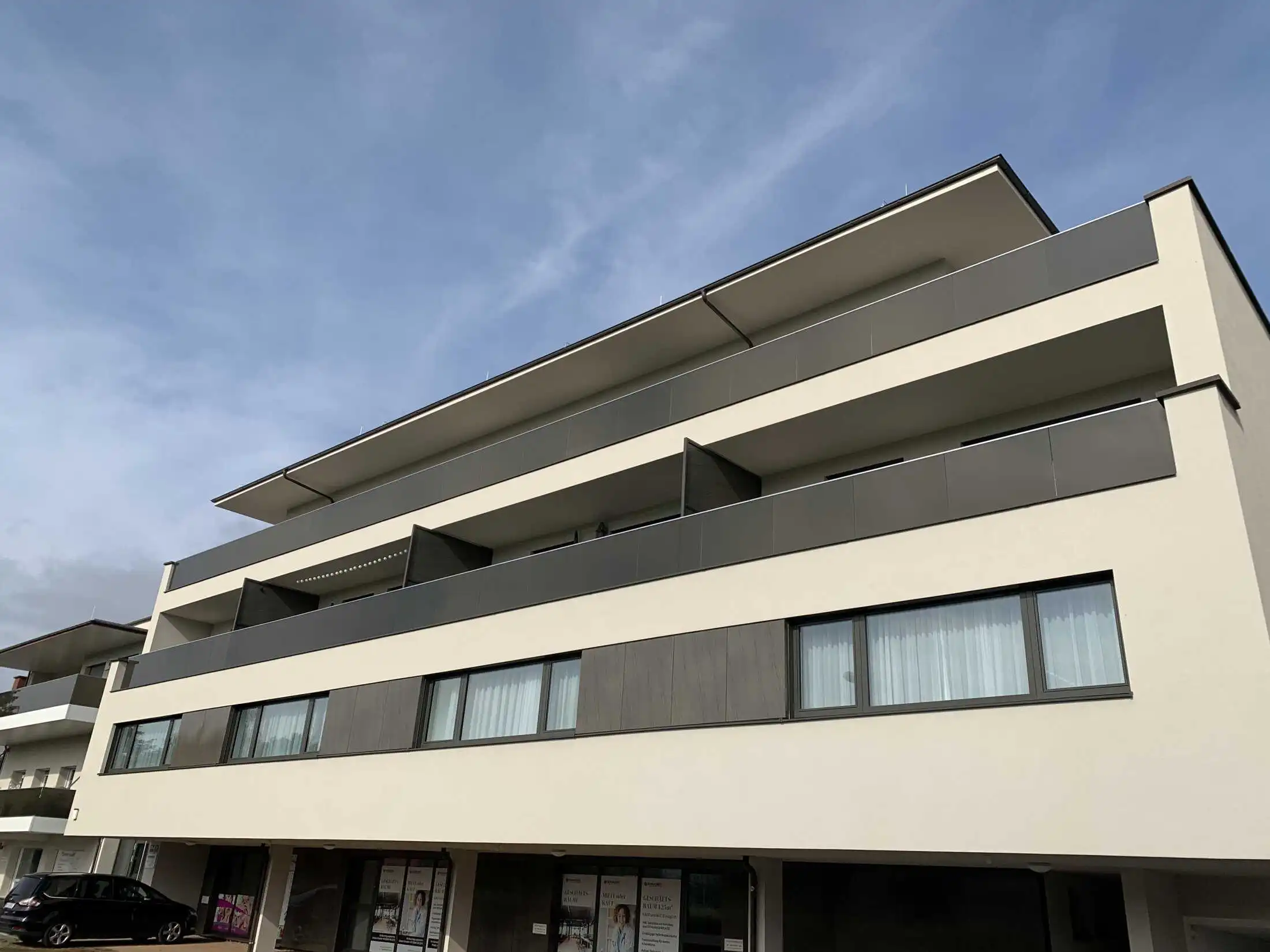 Low-angle view of a modern three-story apartment building with white plaster walls, dark grey balcony panels, and large windows under a blue sky.
