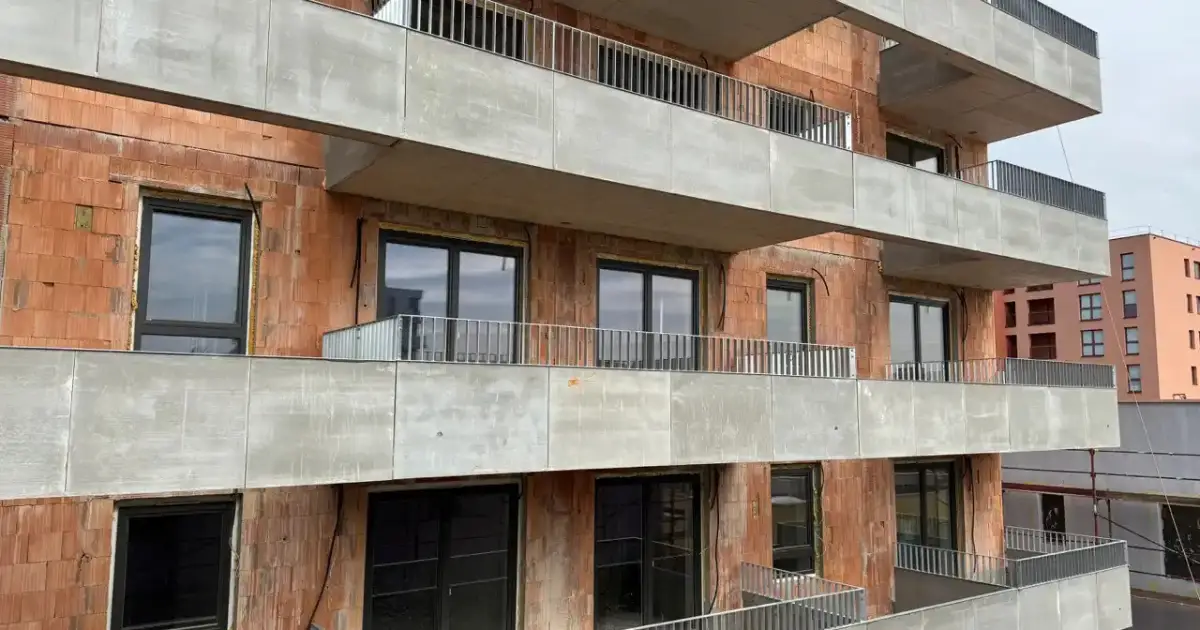 A brick apartment building under construction featuring stacked concrete balconies with metal railings and dark-framed windows.