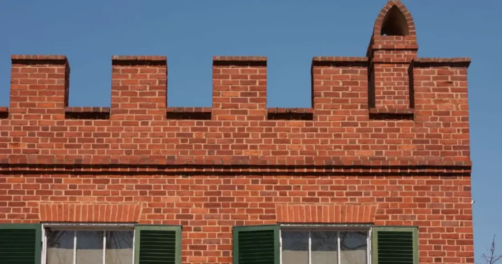 A red brick building featuring a crenelated parapet wall with rectangular notches and a small pointed chimney against a blue sky.
