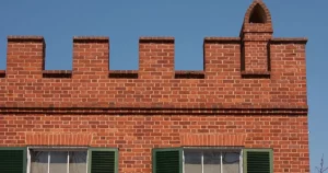 A red brick building featuring a crenelated parapet wall with rectangular notches and a small pointed chimney against a blue sky.