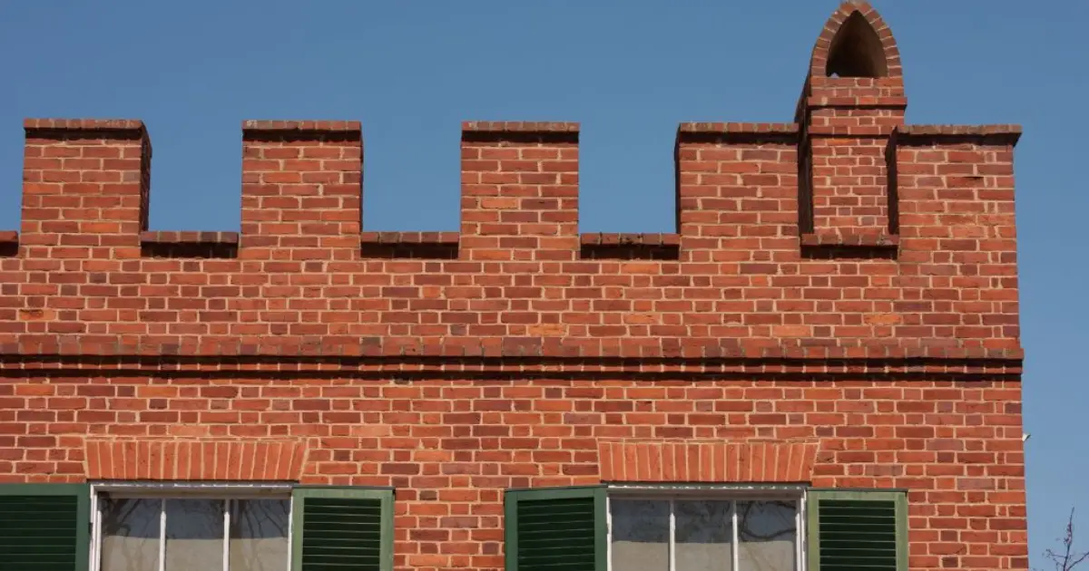 A red brick building featuring a crenelated parapet wall with rectangular notches and a small pointed chimney against a blue sky.