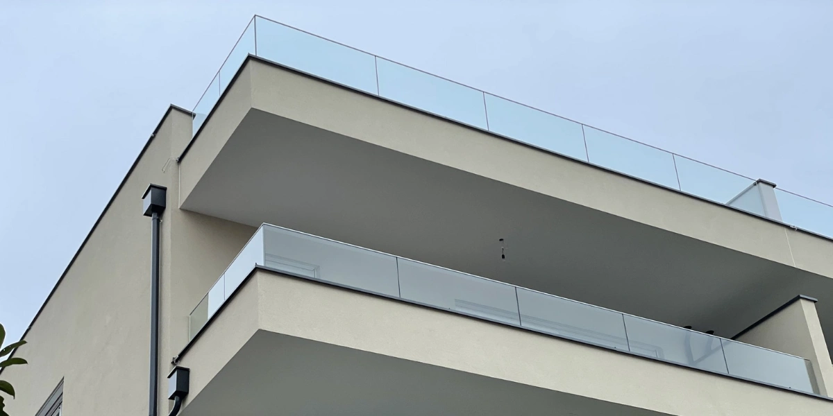 A low-angle shot of two modern, minimalist balconies with sleek glass railings and beige exterior walls under an overcast sky.